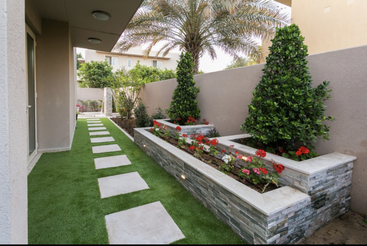 Narrow side yard with artificial turf, square stepping stones and a raised stone planter with flowers